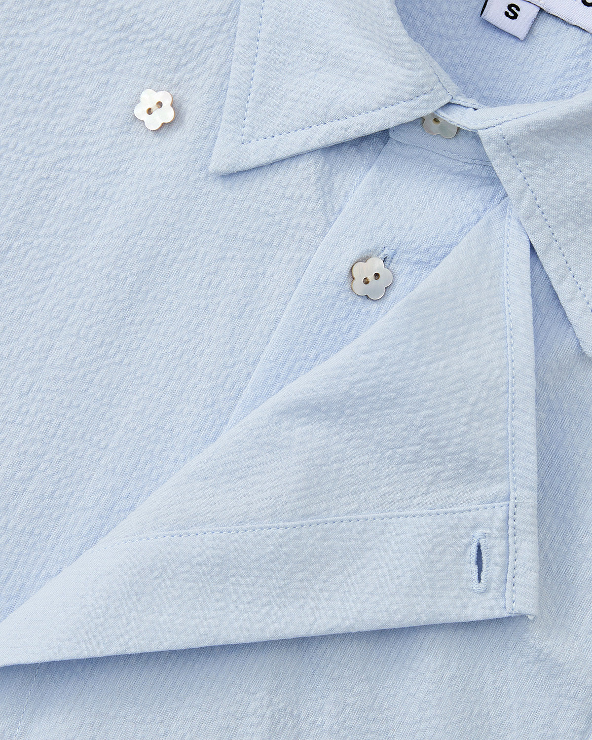 A close-up of the Sundae School SKY BLUE TIE GORUM SHIRT shows its folded collar, off-white flower-shaped buttons, and a visible 'S' tag indicating size. The shirt features light blue seersucker cotton fabric.