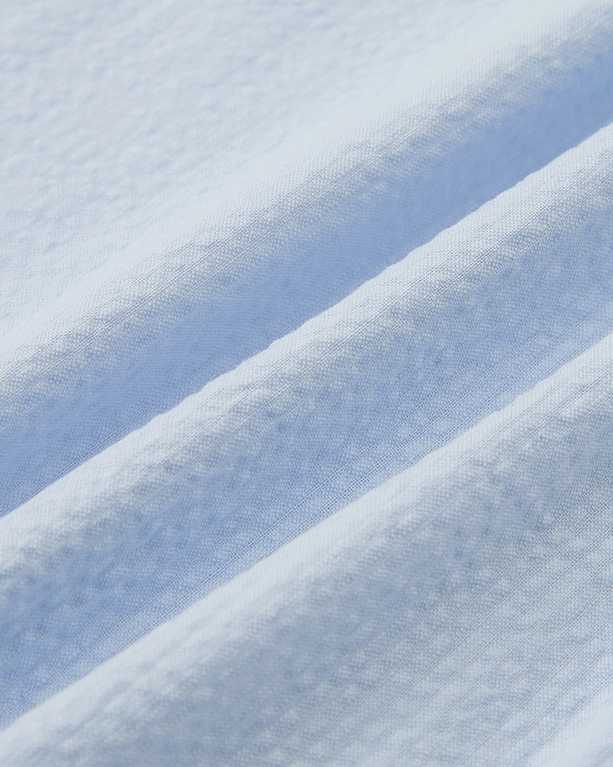 Close-up of the SKY BLUE TIE GORUM SHIRT by Sundae School, featuring light blue seersucker cotton fabric with soft diagonal folds and gentle shadows that showcase its smooth, slightly textured surface.