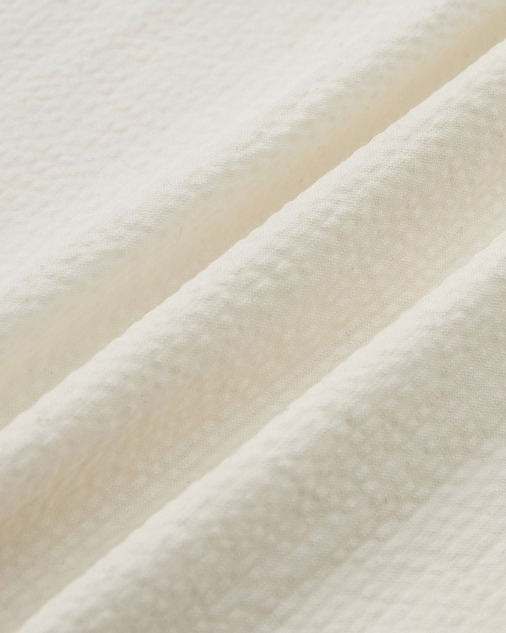 A close-up of the IVORY TIE GORUM SHIRT by Sundae School shows its cream-colored, textured fabric with soft ridges and a subtle woven pattern, reminiscent of seersucker, folded in gentle diagonal lines.