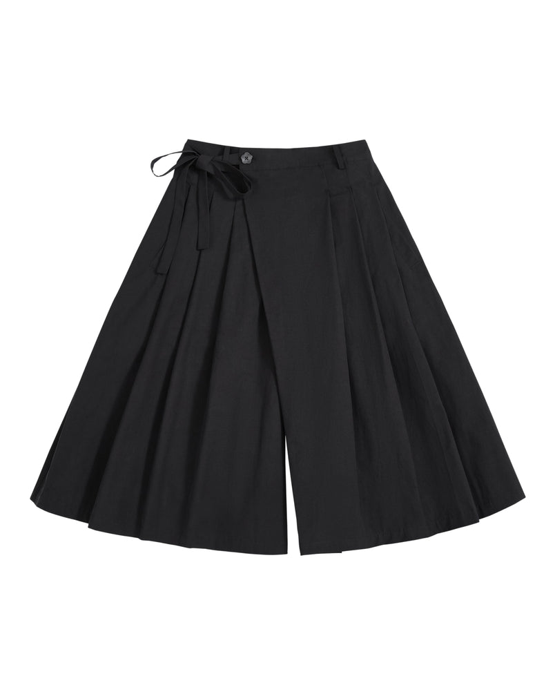The ASYMMETRIC CHIMA CULOTTE by Sundae School, featuring black wide-leg pleated pants with a buttoned waist and a tied ribbon detail on one side, evokes the flowing elegance of a hanbok chima and is displayed laid flat on a white background.
