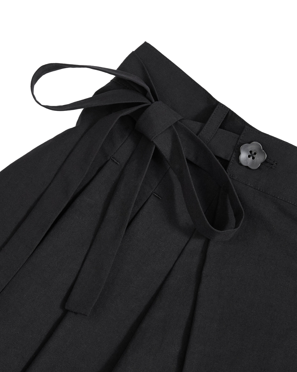 Close-up of the ASYMMETRIC CHIMA CULOTTE by Sundae School, black wrap pants with an asymmetric design, featuring a fabric waist tie in a bow, a flower-shaped black button on the waistband, and pleated detailing below the waistband.