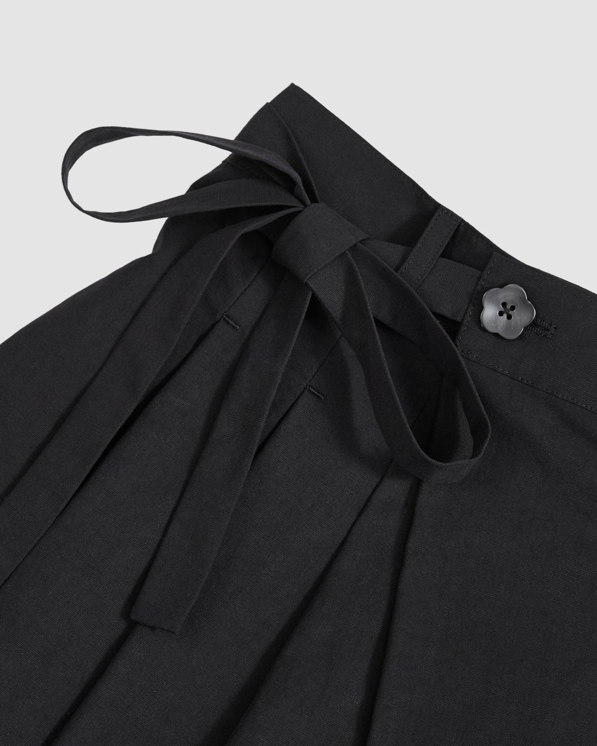 Close-up of the Sundae School BLACK PLEATED HANBOK CULOTTES, featuring pleats, a fabric tie belt knotted in a bow, and a flower-shaped black button at the waistband, shown against a plain light background.