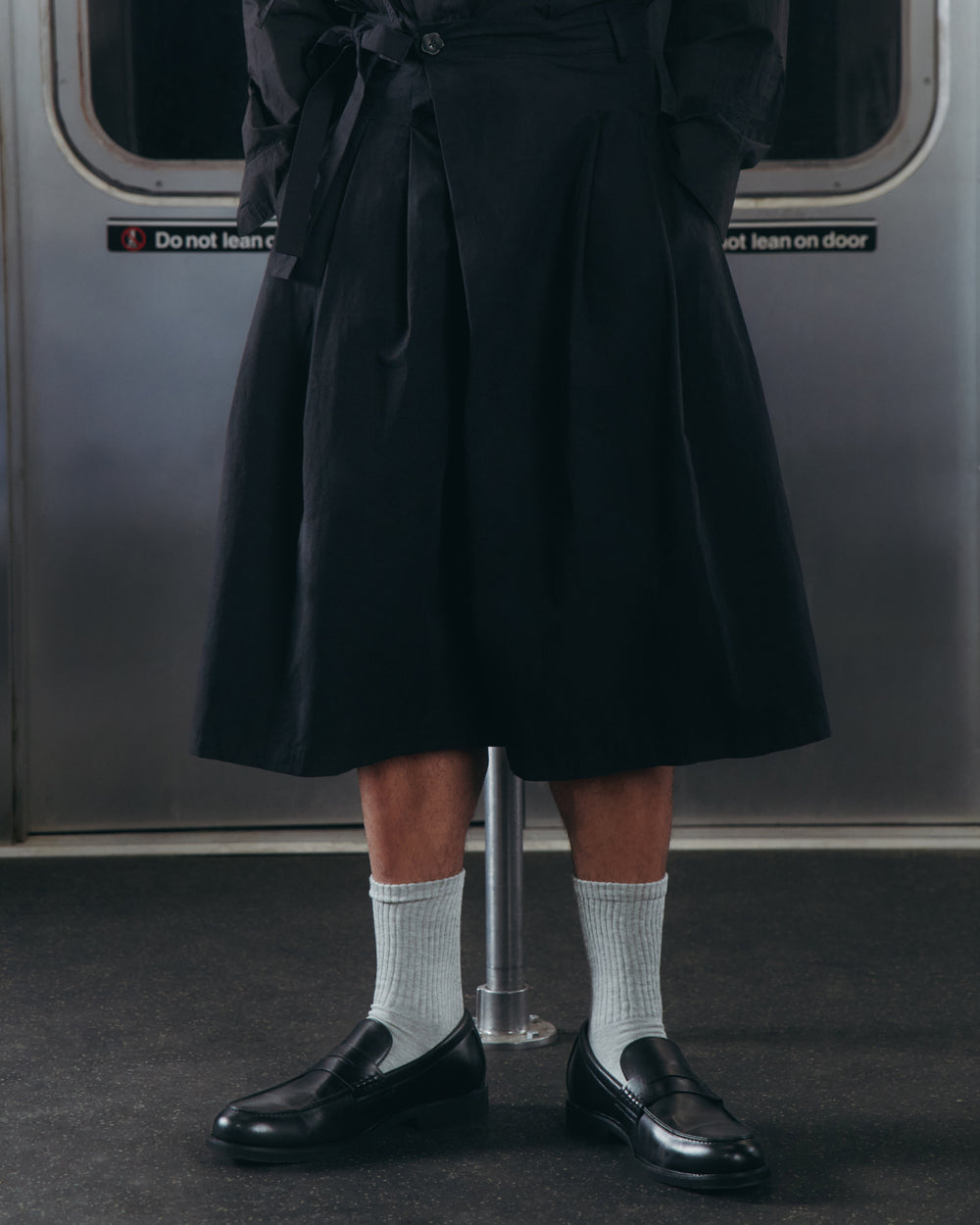 A person wearing the ASYMMETRIC CHIMA CULOTTE by Sundae School, paired with light gray socks and black loafers, stands inside a subway train with metal doors and a “Do not lean on door” sign in the background.
