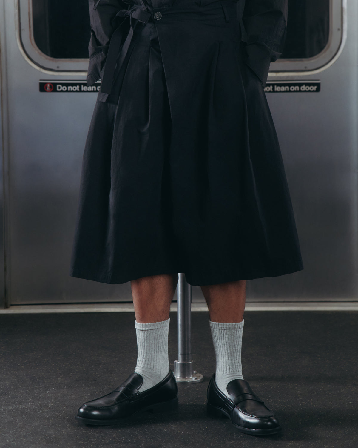 A person wearing the BLACK PLEATED HANBOK CULOTTES by Sundae School, paired with gray socks and black loafers, stands in a subway car with a metal pole and door in the background.