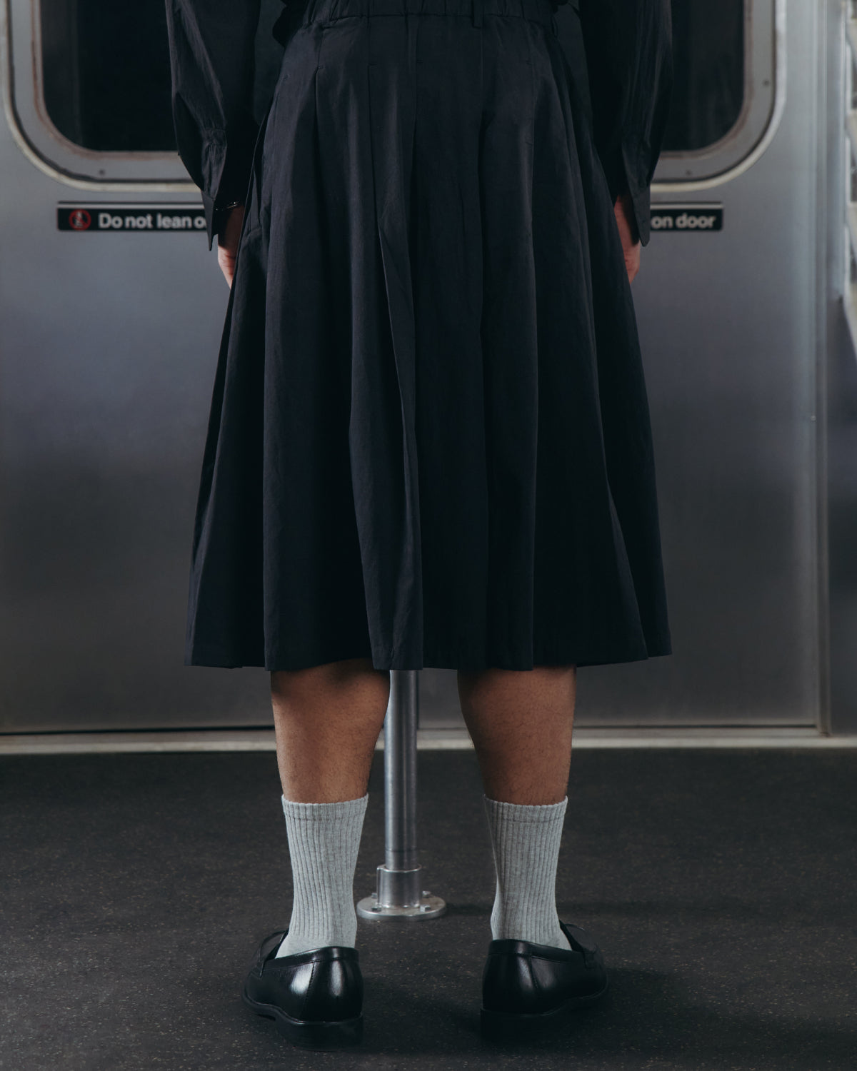 A person stands on a subway, wearing Sundae School's BLACK PLEATED HANBOK CULOTTES, black shoes, and light-colored socks. The subway door and a safety sign can be seen in the background.