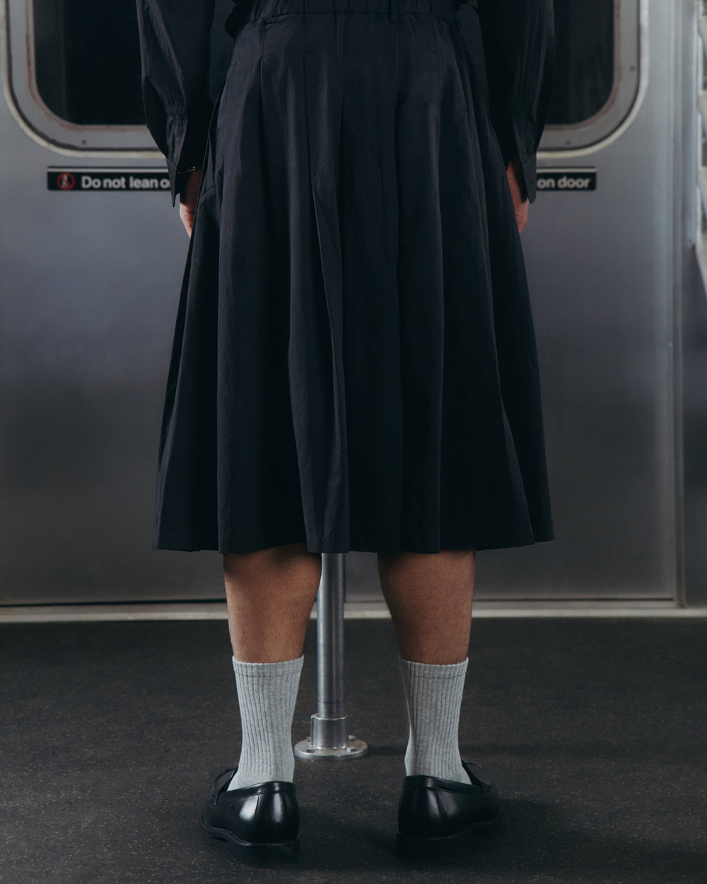 A person stands on a subway train, wearing the Sundae School ASYMMETRIC CHIMA CULOTTE—a flowing black skirt—paired with light gray socks and black shoes. Their back faces the camera, and a metal pole stands in front of them.