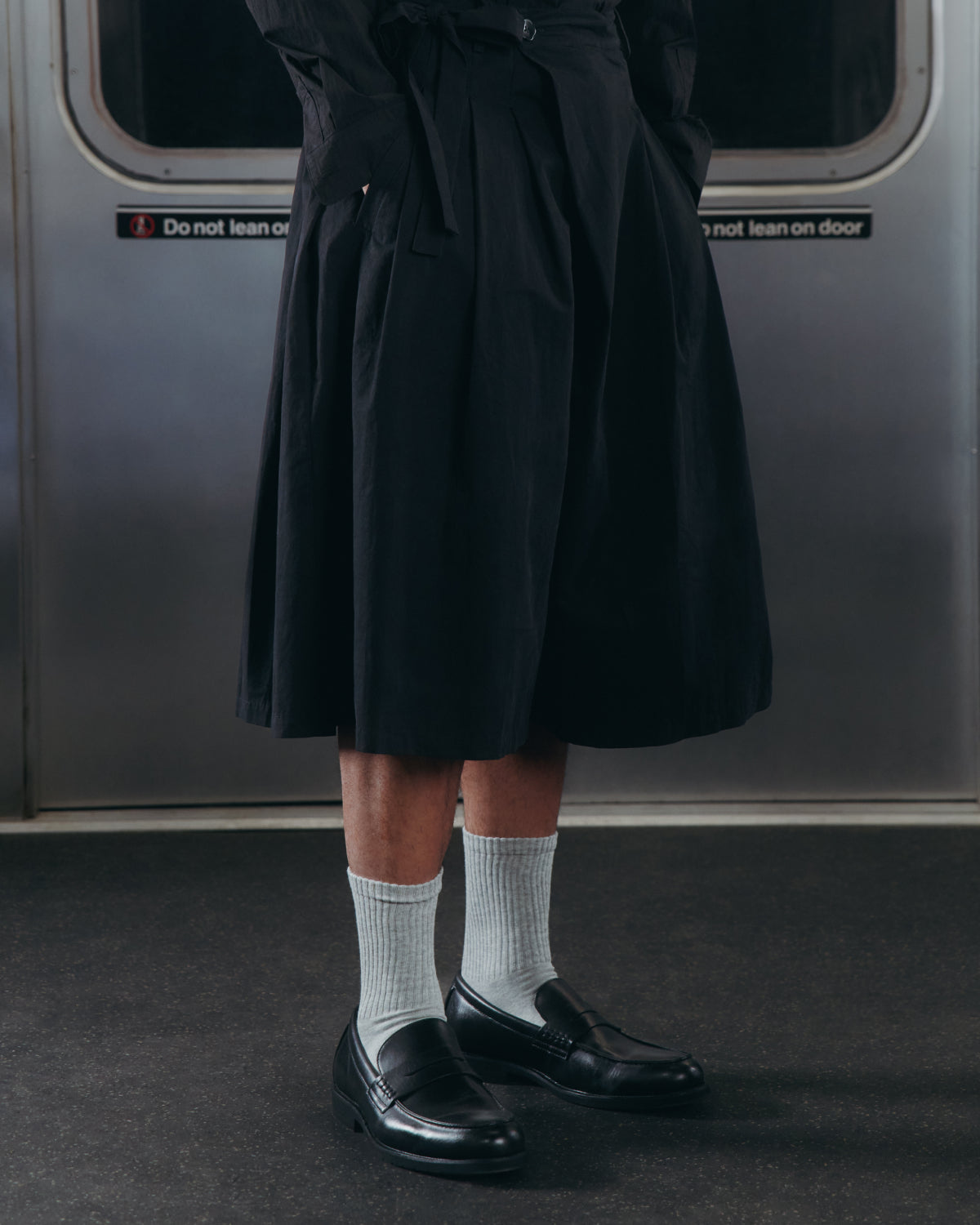 A person stands on a subway train wearing Sundae School's BLACK PLEATED HANBOK CULOTTES, paired with white socks and black loafers. In the background, a subway door displays a “Do not lean on door” sign.