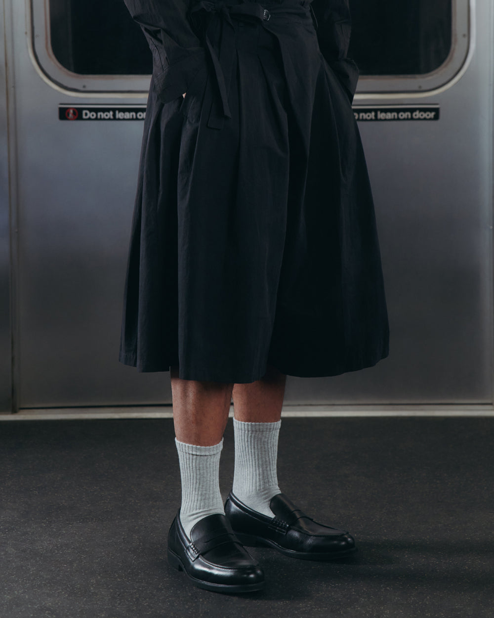 A person wearing the Sundae School ASYMMETRIC CHIMA CULOTTE, paired with white socks and black loafers, stands inside a subway train. Only their lower body is visible. A sign on the train door reads “Do not lean on door.”.