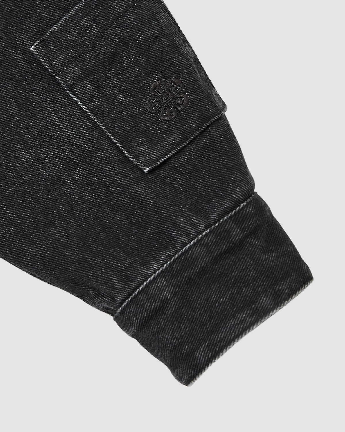 Close-up of a black denim sleeve from the Sundae School BLACK ASYMMETRIC WORK JACKET, showing a flap pocket with a stitched circular emblem. The fabric is slightly faded, set against a plain light gray background.