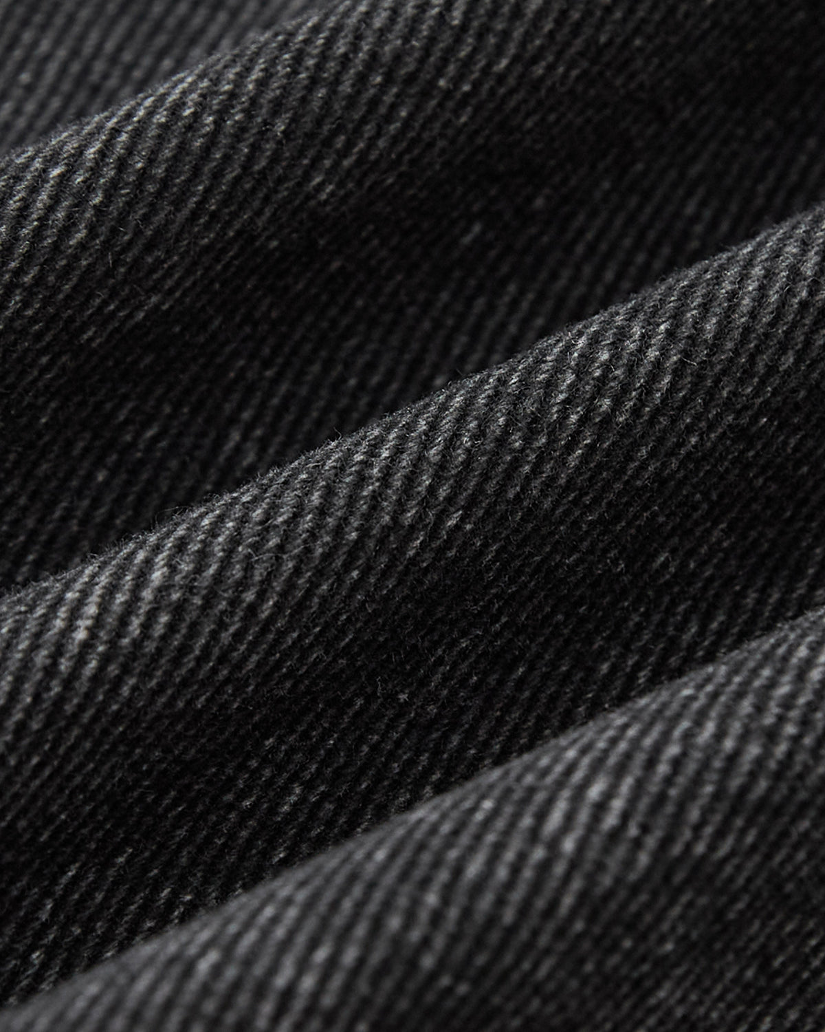 Close-up of the BLACK ASYMMETRIC WORK JACKET by Sundae School, highlighting its black denim fabric with a diagonal weave pattern, visible texture, and soft folds.