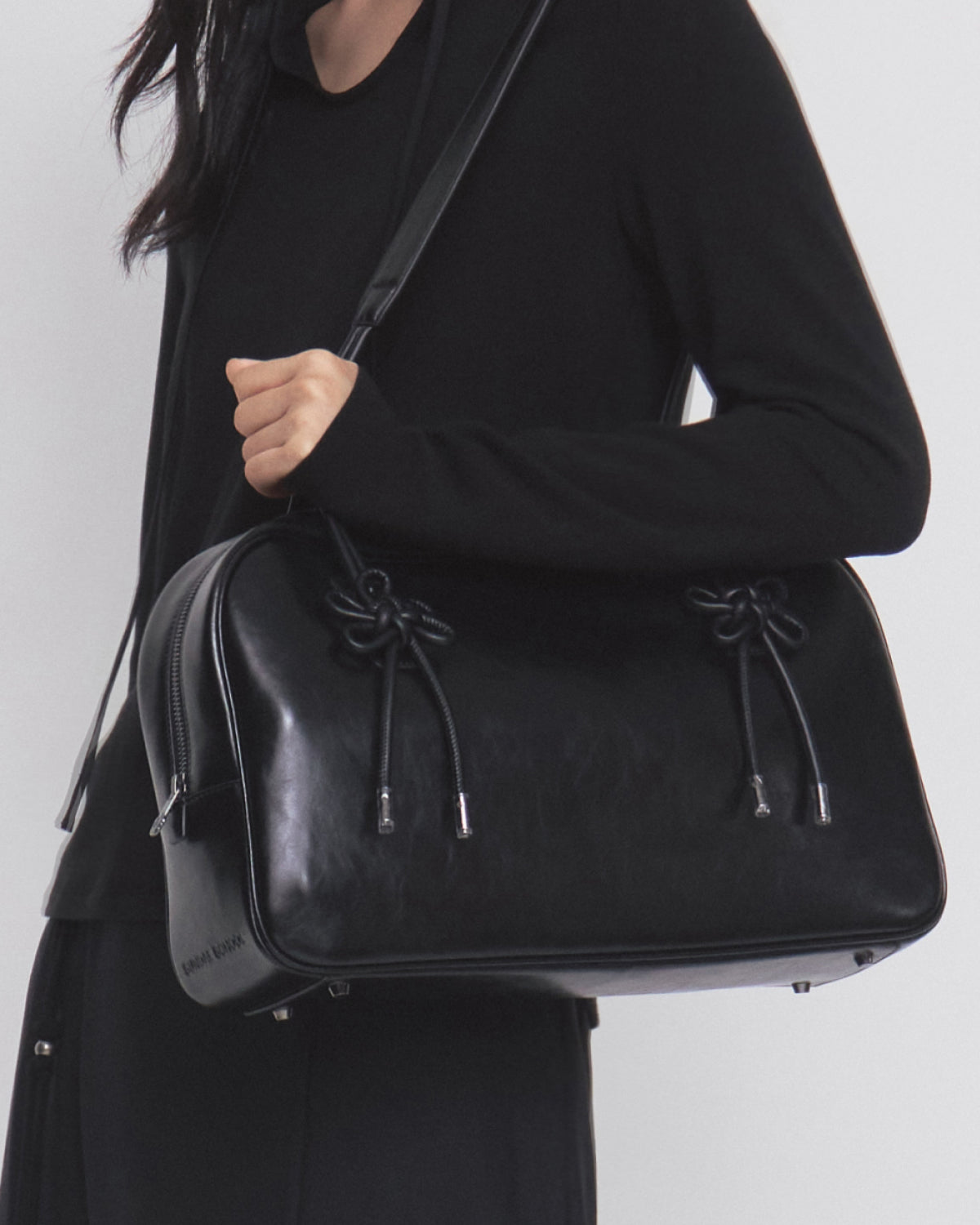 A person wearing a black outfit holds the Sundae School BLACK AUDREY BOWLING BAG, a large black leather handbag featuring decorative knotted details and metal feet, slung over their shoulder.