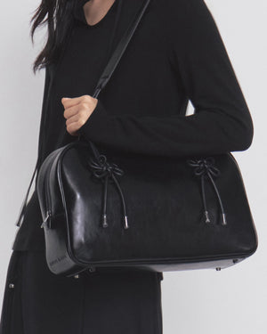 A person wearing a black outfit holds the Sundae School BLACK AUDREY BOWLING BAG, a large black leather handbag featuring decorative knotted details and metal feet, slung over their shoulder.