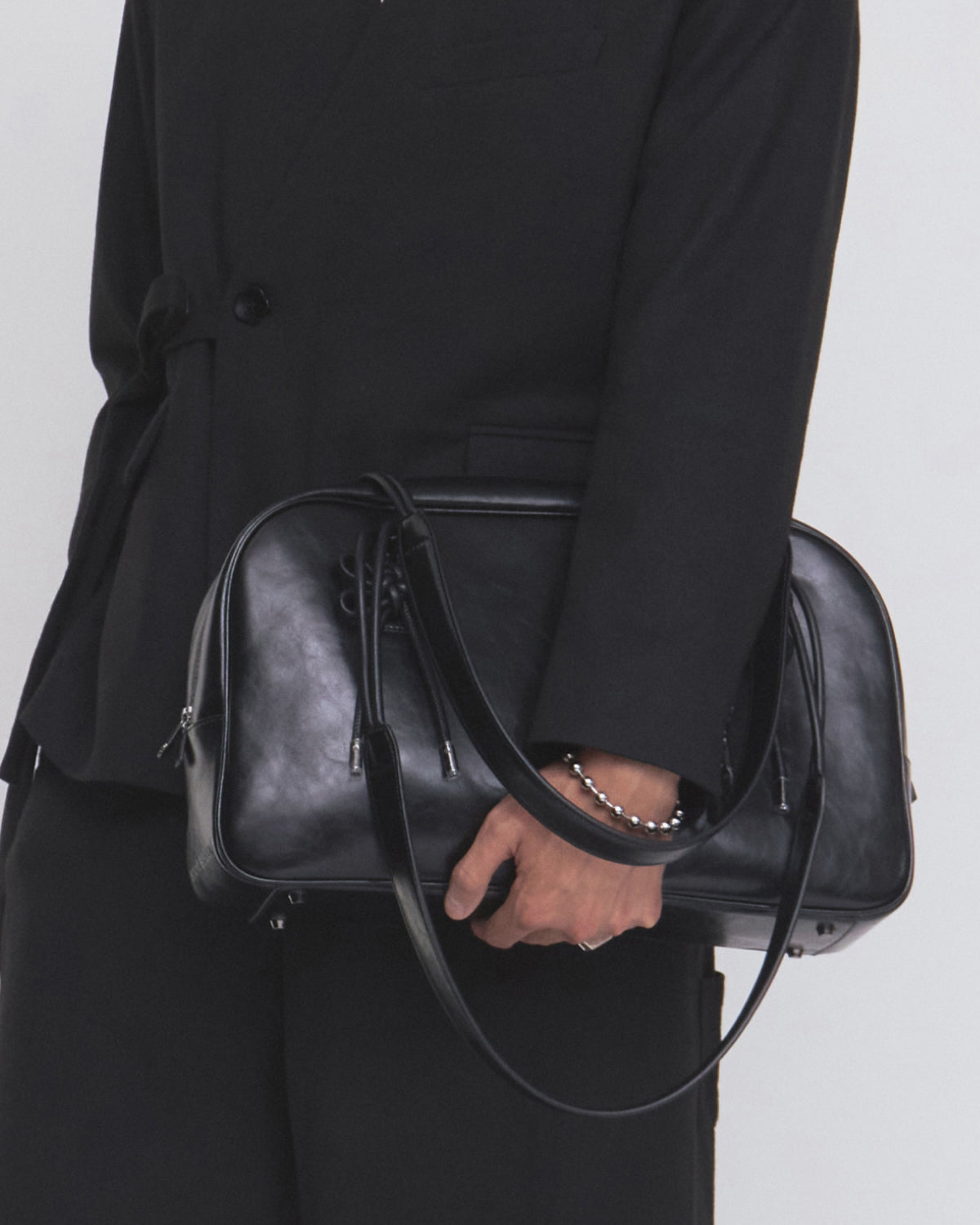 A person in a black suit holds the Sundae School BLACK AUDREY BOWLING BAG, a large black leather handbag with long handles, shown from the shoulders down against a plain background.
