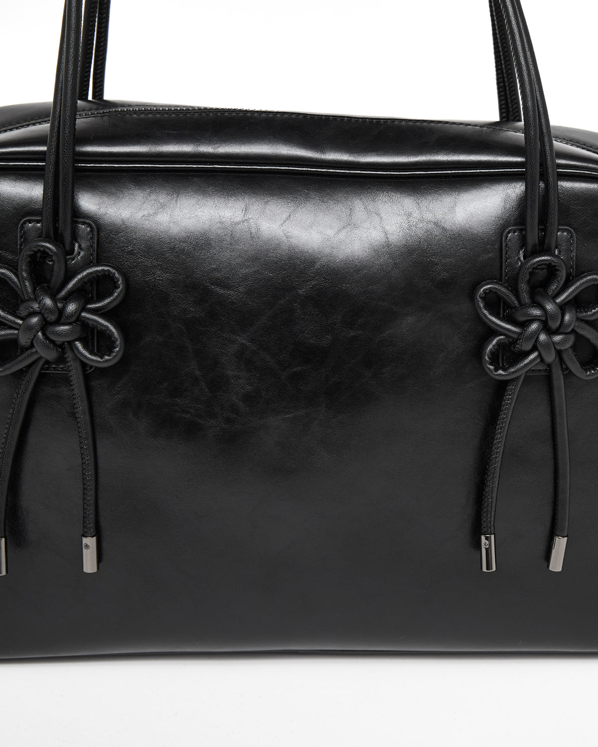 A close-up of the Sundae School BLACK AUDREY BOWLING BAG, featuring shiny black leather, two black knotted floral details, and metal-tipped drawstrings on each side of the handle bases.