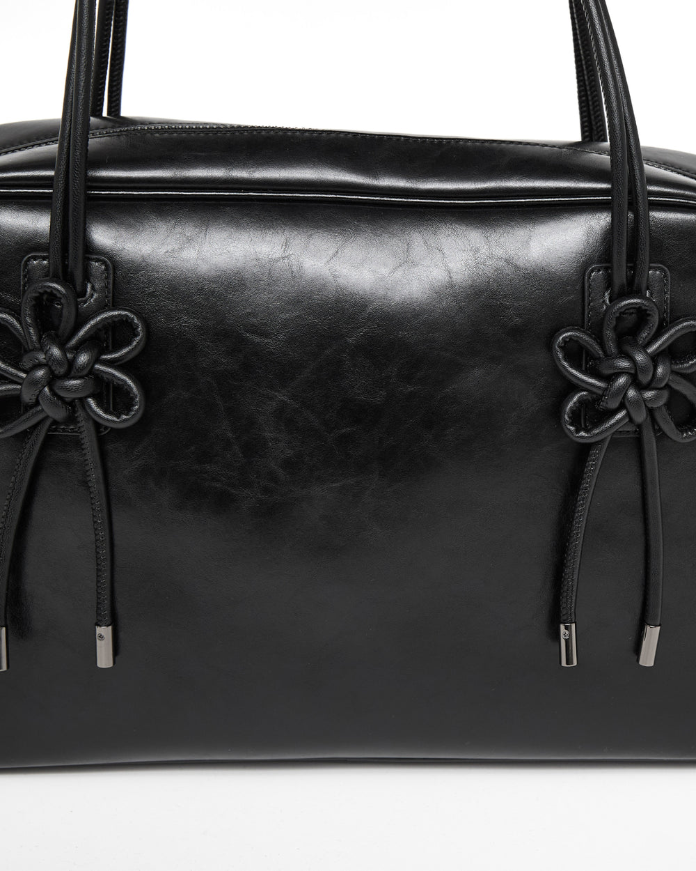 A close-up of the Sundae School BLACK AUDREY BOWLING BAG, featuring shiny black leather, two black knotted floral details, and metal-tipped drawstrings on each side of the handle bases.