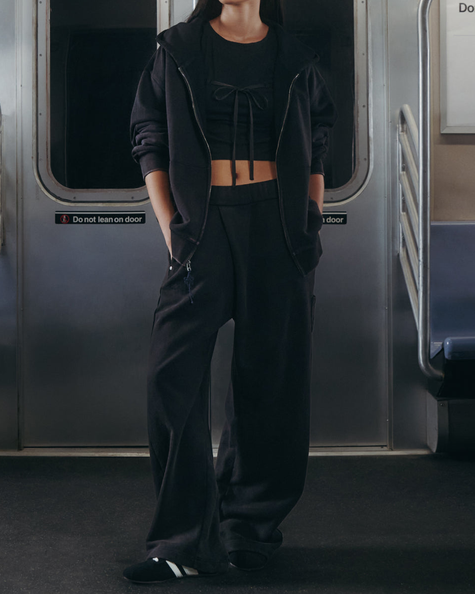 A person stands in a subway car wearing Sundae School’s BLACK WASH WRAP SWEAT BAJI. Their hands are in their pockets, and the subway seats around them are empty.