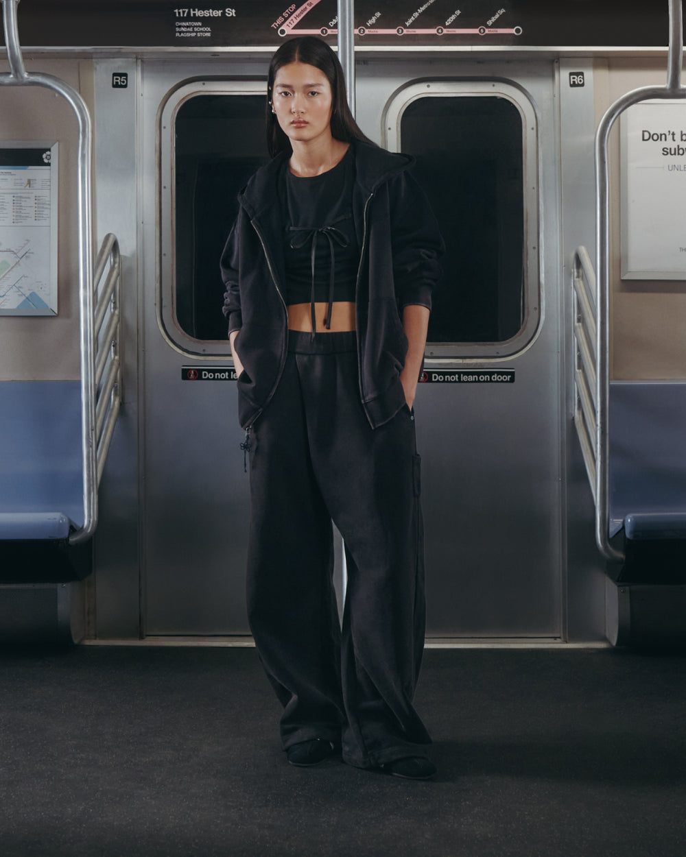 A person stands in the middle of a subway car wearing the BLACK TIE GORUM BABY TEE by Sundae School, paired with loose black pants and a black jacket. Subway seats and maps are visible in the background, hands tucked in pockets.