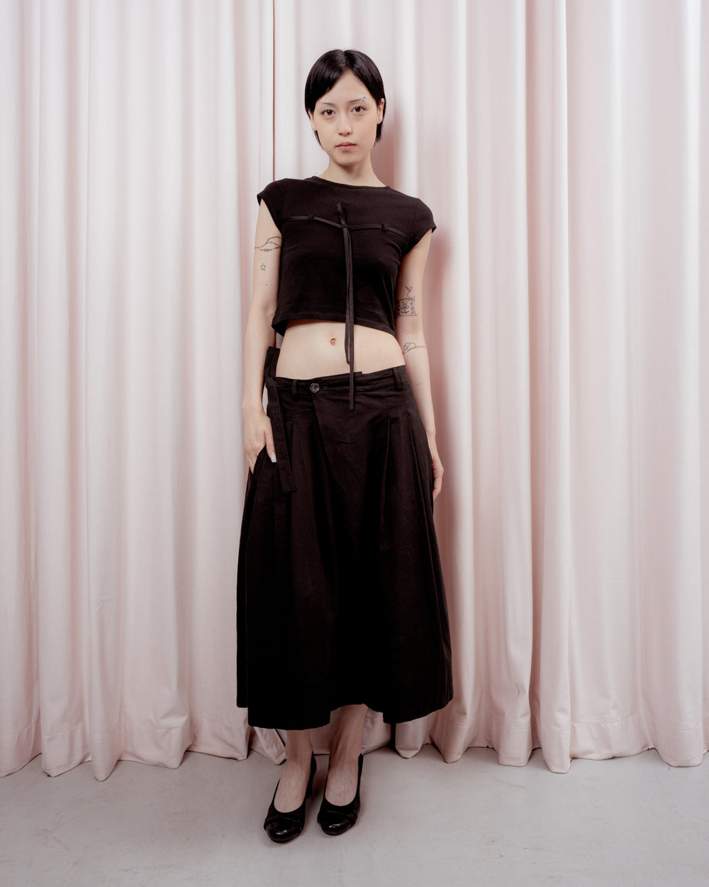 Full-length front view of a person with short black hair standing against a light pink curtain backdrop. They are wearing a cropped black short-sleeved t-shirt with a thin tie-detail at the chest, a low-waisted flowing black midi skirt, and black low-heeled shoes.