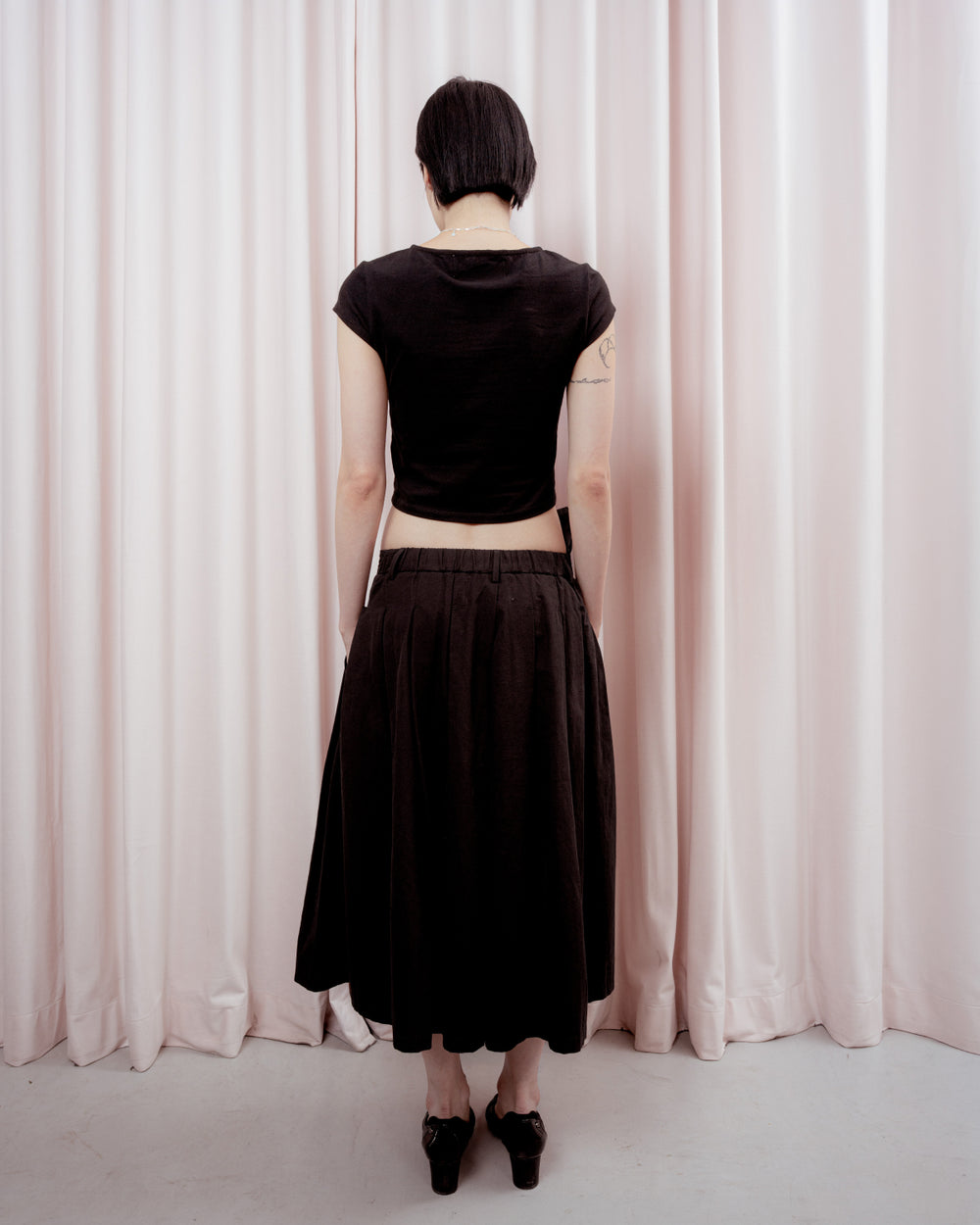 Full-length back view of a person with short black hair standing against a light pink curtain backdrop. They are wearing a cropped black short-sleeved t-shirt and a loose black midi skirt, revealing their lower back and arms. They are wearing black low-heeled shoes.