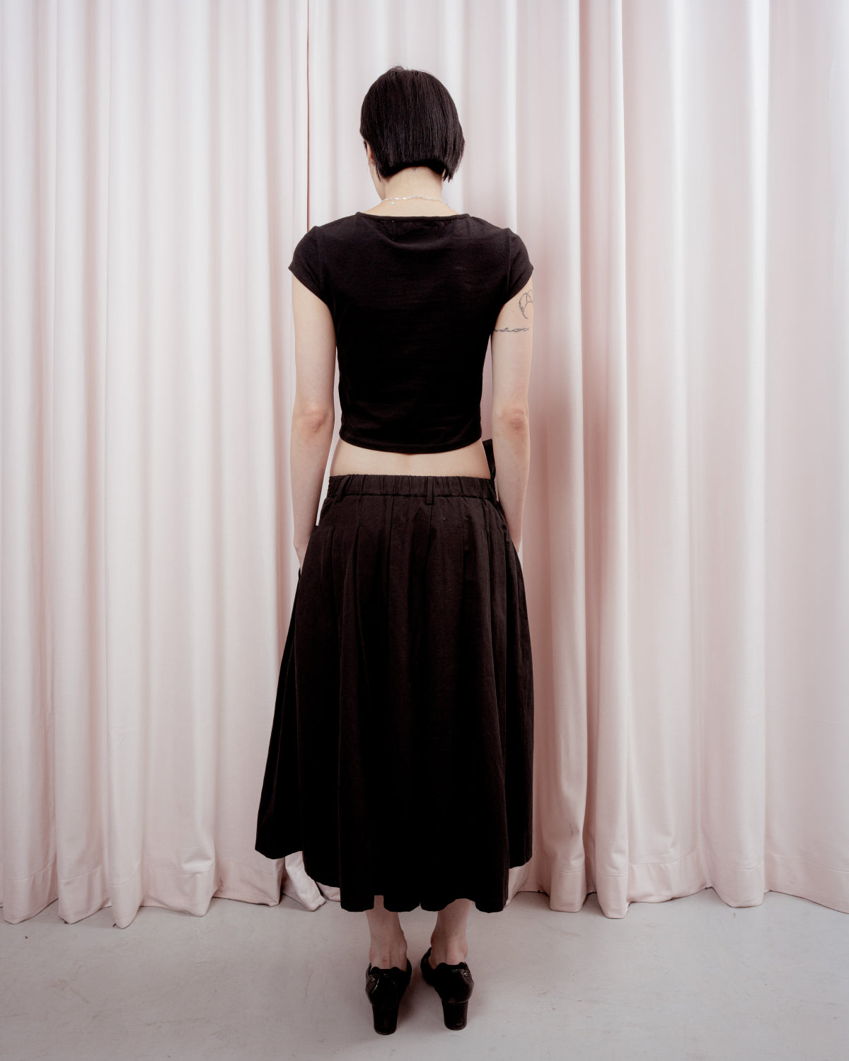 Full-length back view of a person with short black hair standing against a light pink curtain backdrop. They are wearing a cropped black short-sleeved t-shirt and a loose black midi skirt, revealing their lower back and arms. They are wearing black low-heeled shoes.