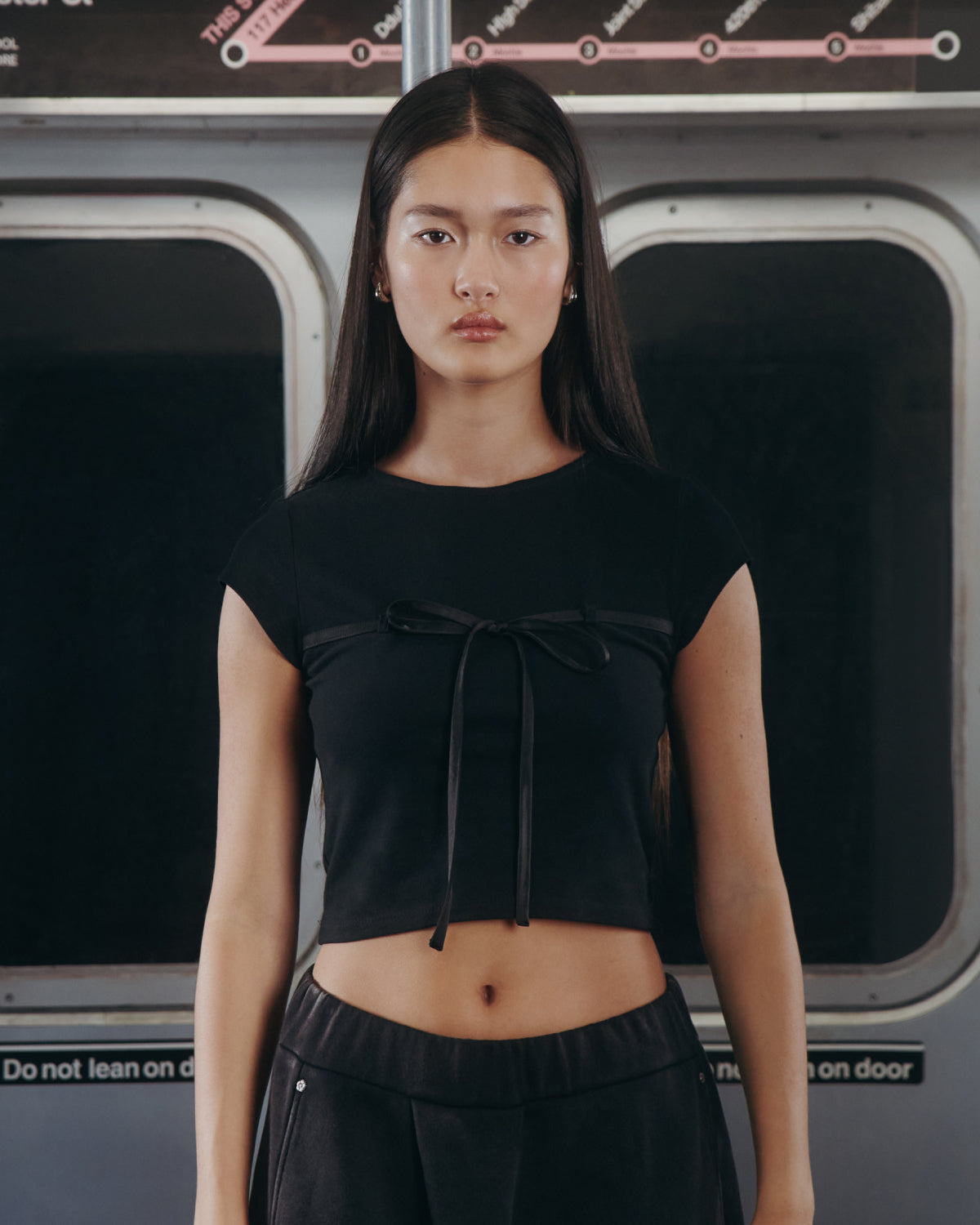 A woman with long dark hair wears the Sundae School BLACK TIE GORUM BABY TEE and black pants, standing by a subway window beneath "Do not lean on door" signs in a dimly lit setting.