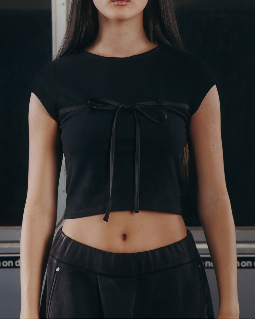 A woman with long dark hair wears the Sundae School BLACK TIE GORUM BABY TEE and black pants, standing by a subway window beneath "Do not lean on door" signs in a dimly lit setting.