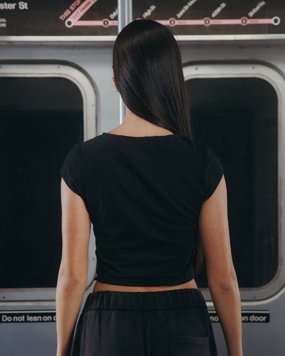 A person with long straight hair, wearing the BLACK TIE GORUM BABY TEE by Sundae School and black pants, stands with their back to the camera inside a subway train, where a map and signs are visible above the windows.