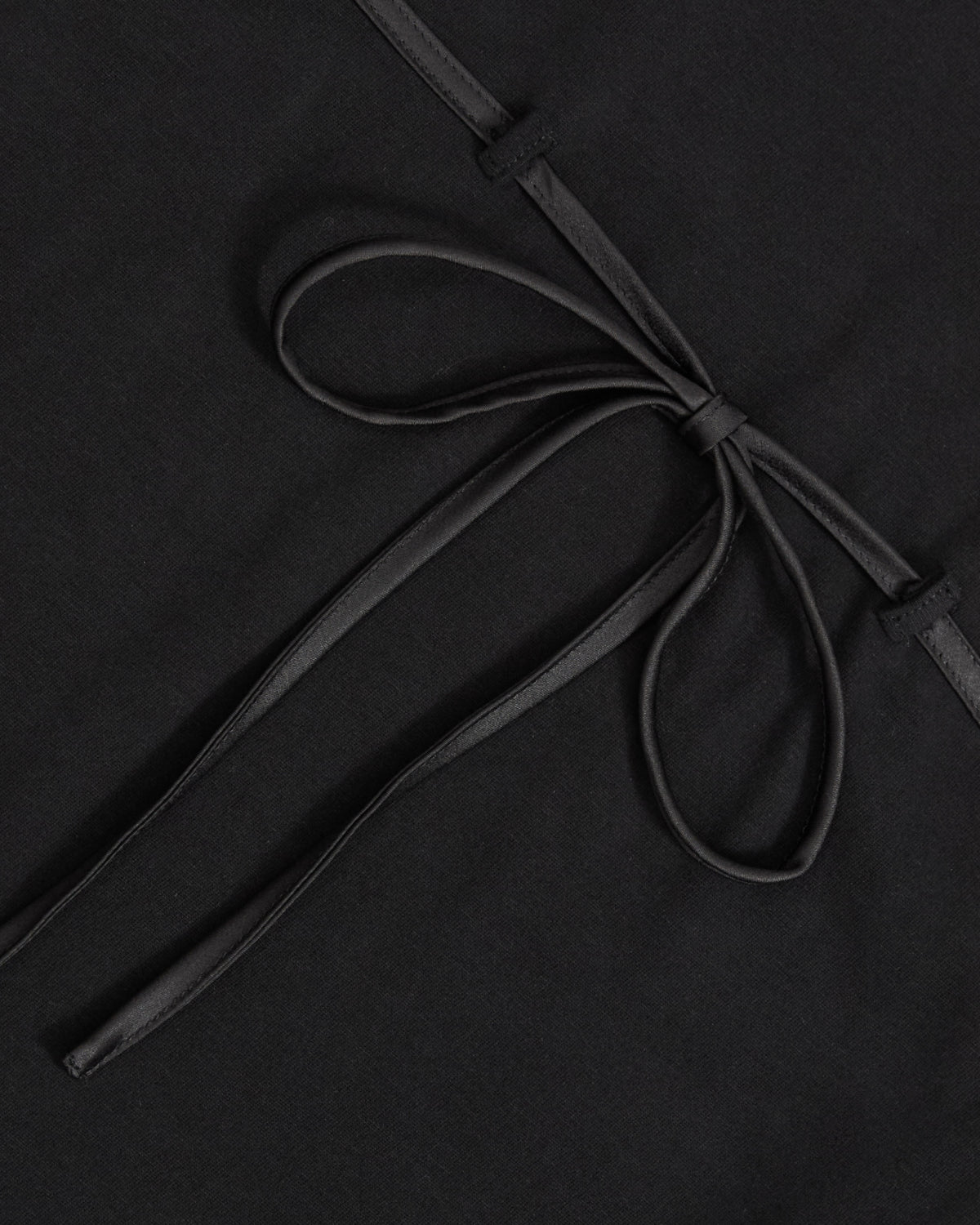 A close-up of the Sundae School BLACK TIE GORUM BABY TEE showcases a neatly tied, thin black ribbon bow set against a smooth, matte black fabric background. The elegant bow highlights the modern, minimalist design of this form-fitting women’s shirt.
