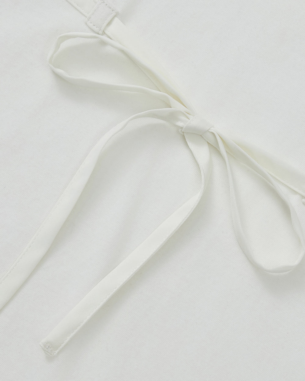 Close-up of the WHITE TIE GORUM BABY TEE by Sundae School, featuring a neat chest strap bow tied from its waist straps, lying flat on a white surface.