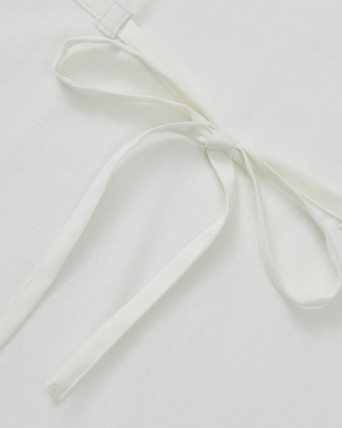 Close-up of the WHITE TIE GORUM BABY TEE by Sundae School, featuring a neat chest strap bow tied from its waist straps, lying flat on a white surface.