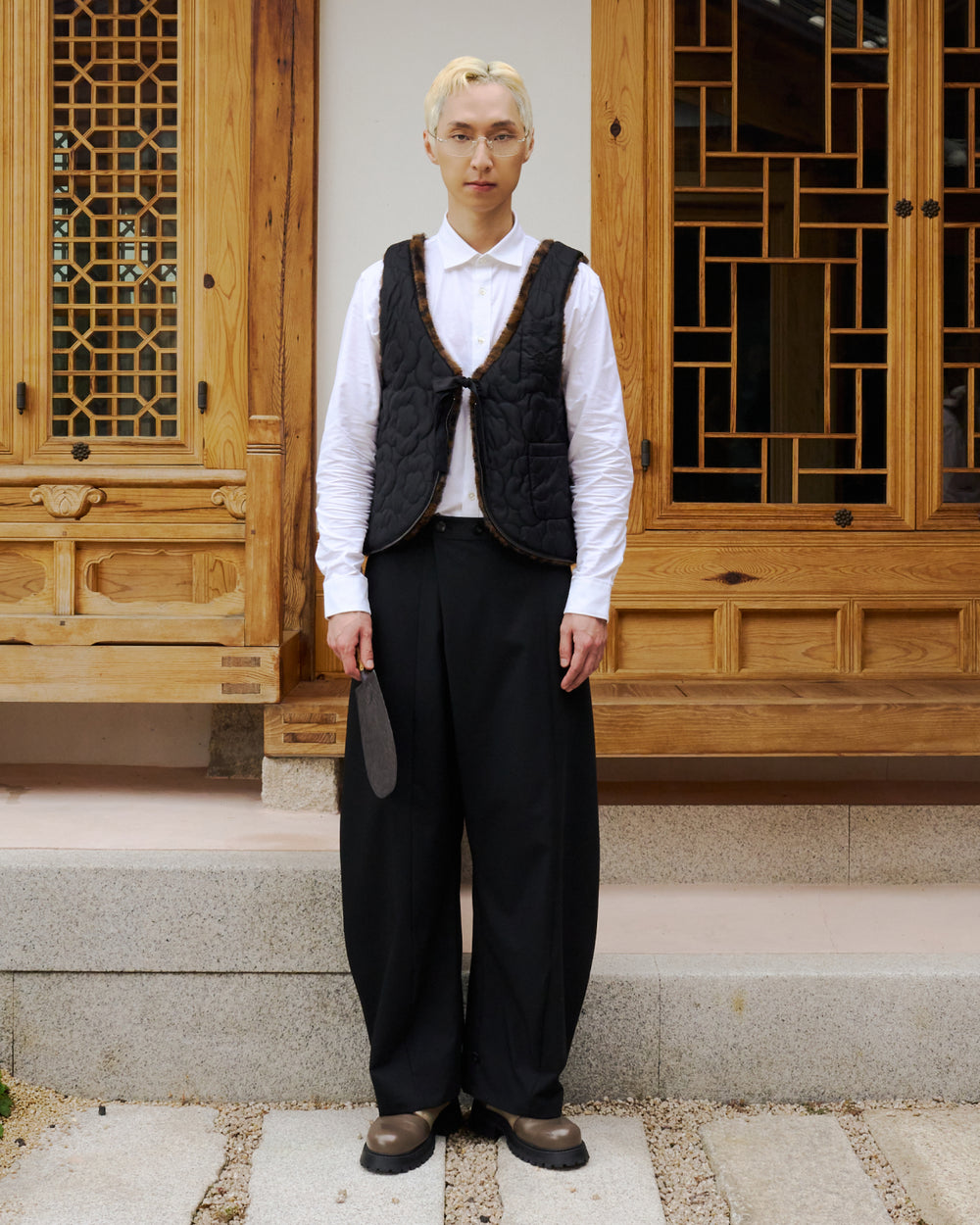 A person with short blond hair, glasses, and light skin stands on stone steps in front of a wooden building with decorative lattice windows. They wear a white shirt paired with the BLACK BAEJA VEST / BAG from Sundae School, wide black pants, and brown shoes, holding a fan.