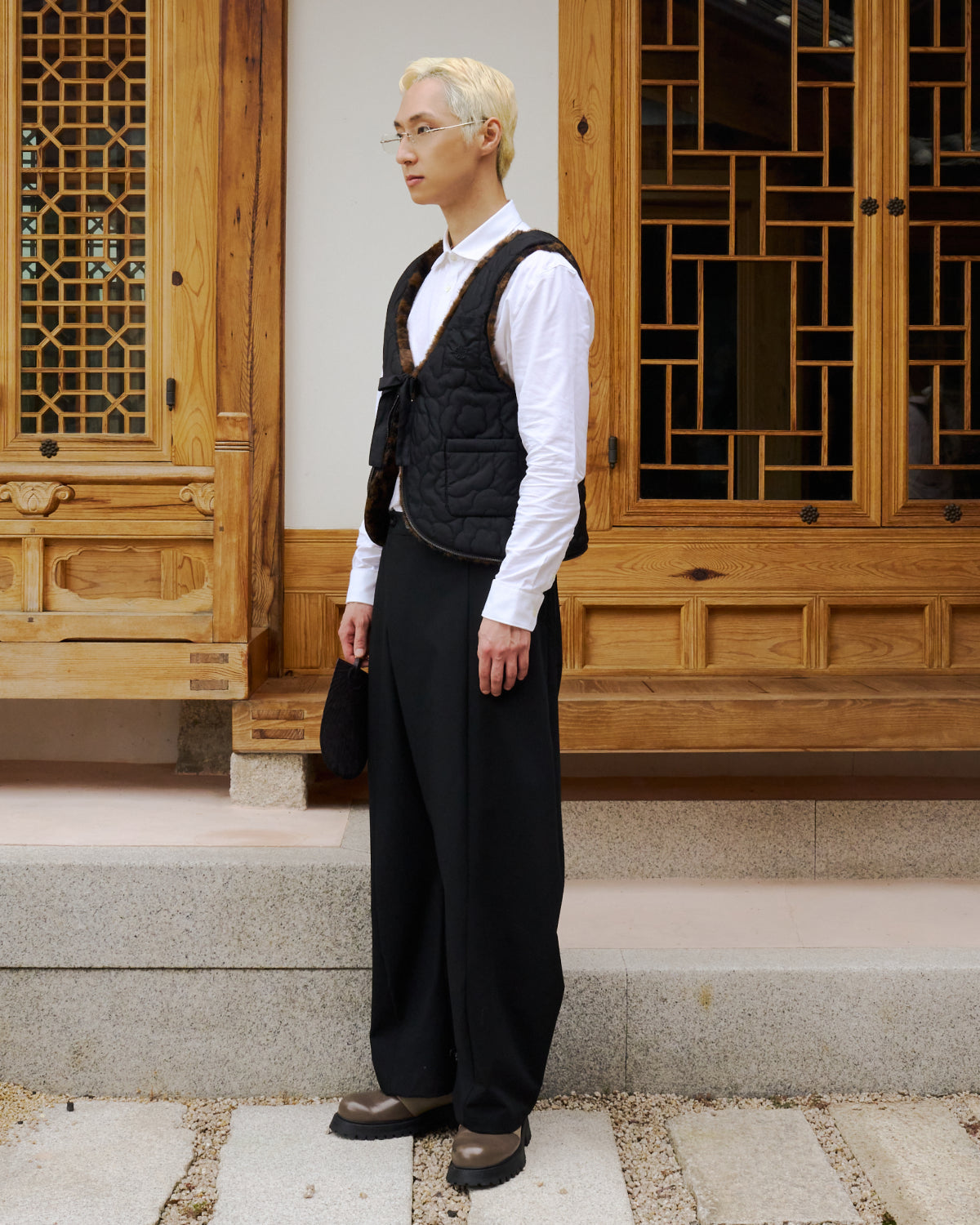 A person with short blond hair, glasses, and dressed in a white shirt, Sundae School’s BLACK BAEJA VEST / BAG layered over wide black pants and brown shoes stands on stone steps in front of a traditional wooden building.