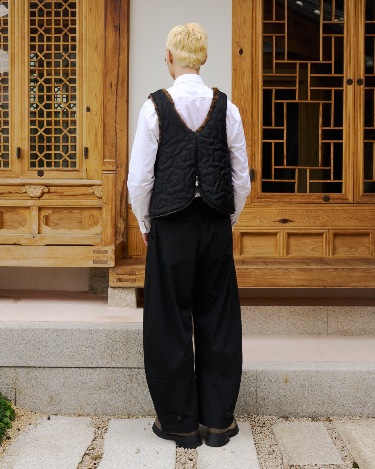 A person with short blond hair, dressed in a white shirt and Sundae School’s BLACK BAEJA VEST / BAG, paired with wide black pants, stands facing away in front of a traditional wooden building adorned with decorative windows.