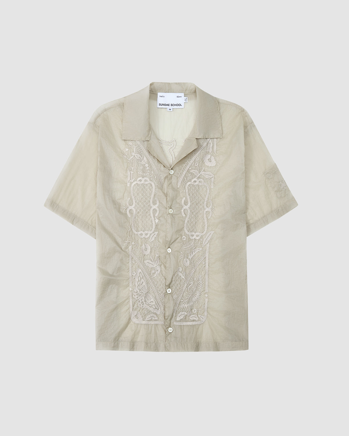 The BARONG SHIRT by Sundae School is a light beige, short-sleeve button-up with a collar and white embroidery on the front. Inspired by the Barong Tagalog, it offers a relaxed fit and casual style.