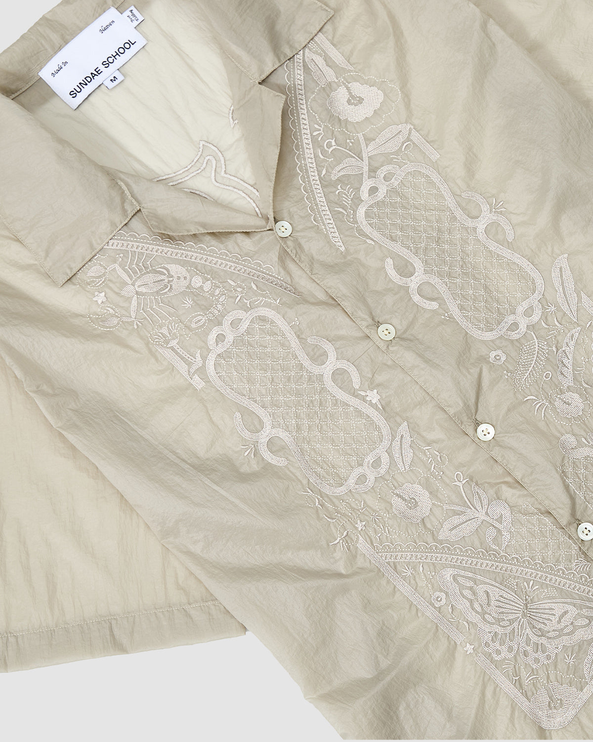The BARONG SHIRT by Sundae School is a beige, short-sleeved shirt inspired by the Barong Tagalog, featuring white floral and geometric embroidery on the front, a collar with white buttons, and a Sundae School label inside.