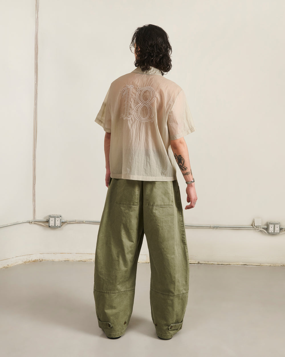 In a minimalist room, a person with shoulder-length wavy hair and left arm tattoos wears Sundae School's BARONG SHIRT—nylon, translucent, "18" on the back—paired with loose olive pants, creating a striking Filipino silhouette.
