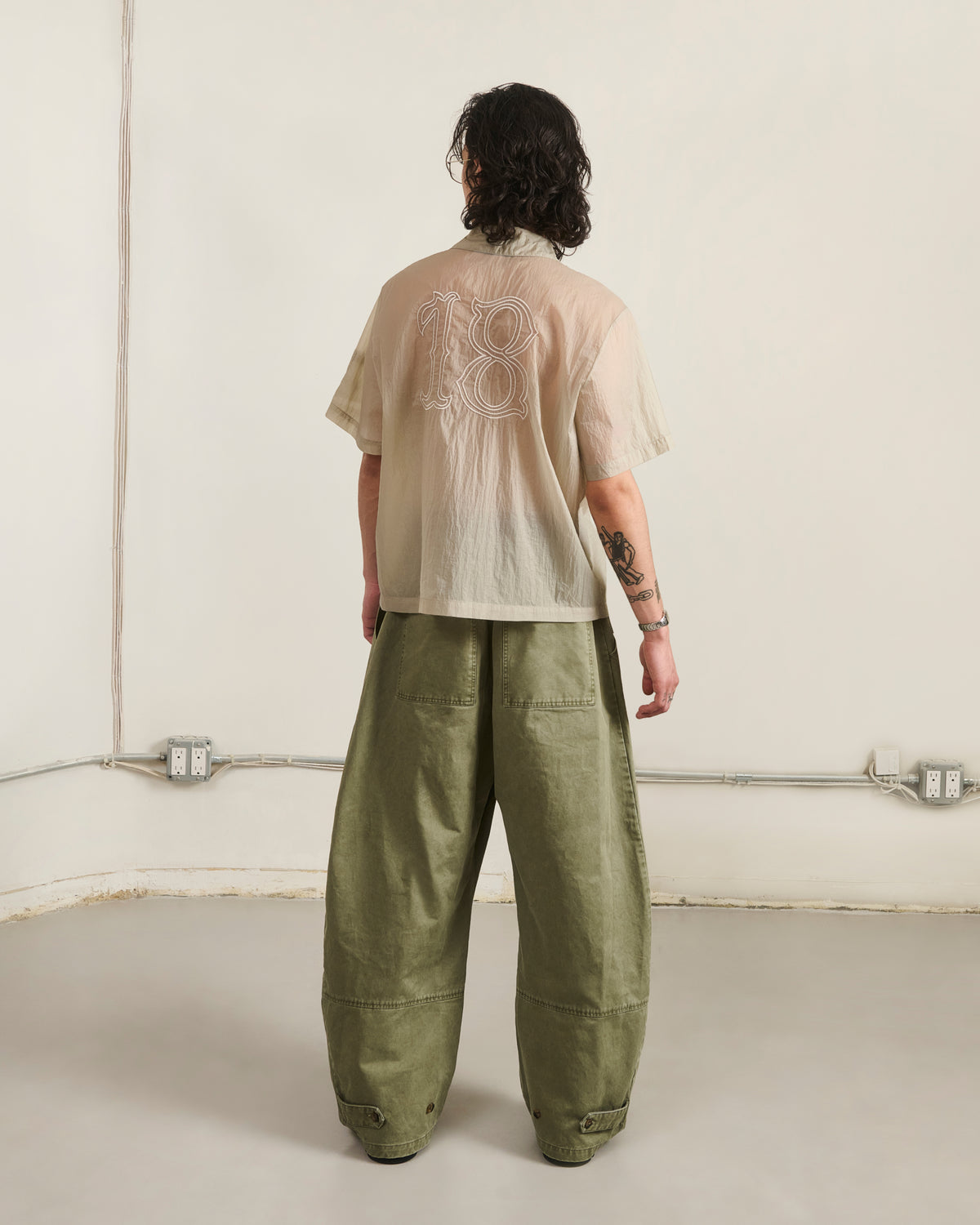 In a minimalist room, a person with shoulder-length wavy hair and left arm tattoos wears Sundae School's BARONG SHIRT—nylon, translucent, "18" on the back—paired with loose olive pants, creating a striking Filipino silhouette.