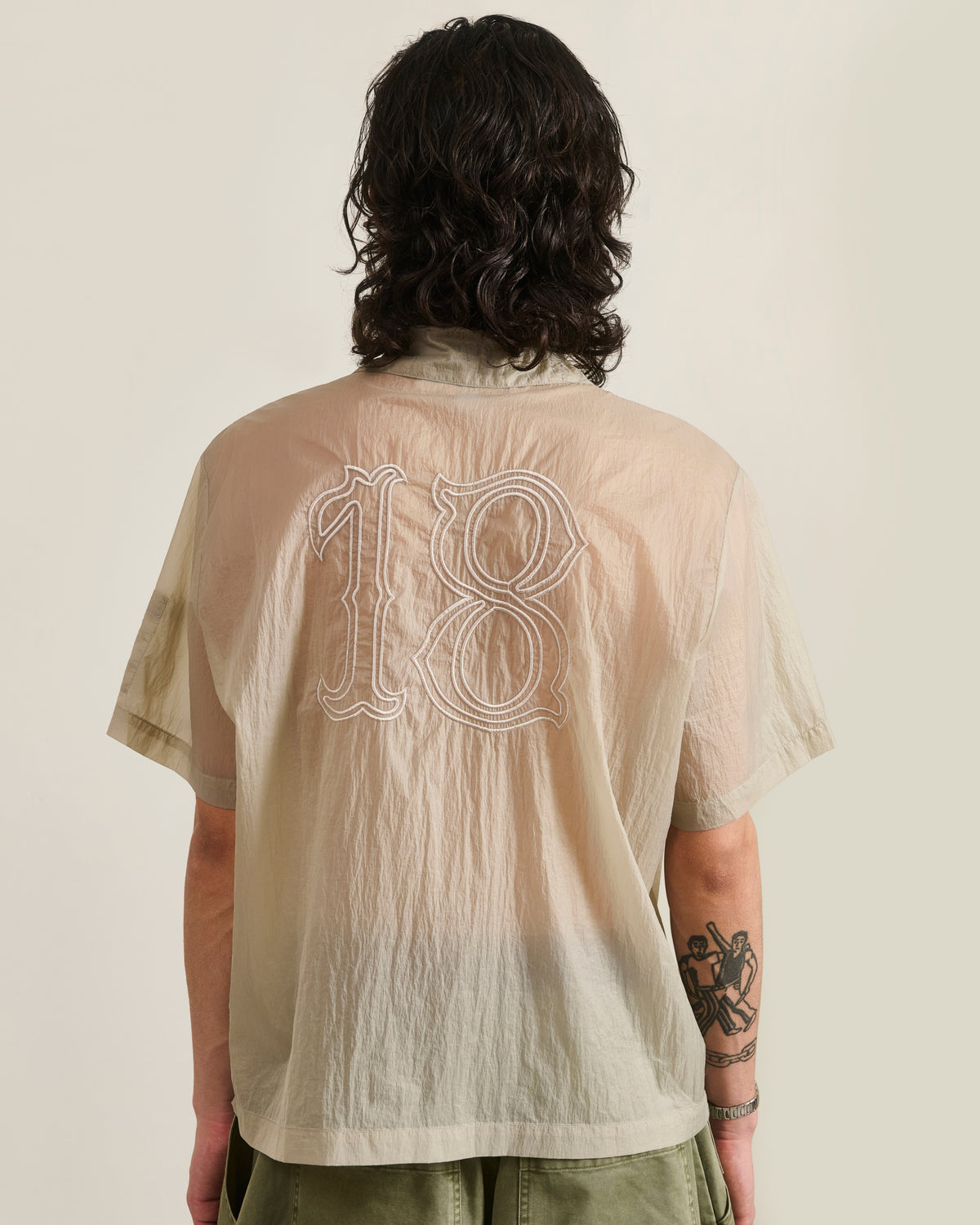 A person with long dark hair, wearing the Sundae School BARONG SHIRT—a sheer beige nylon shirt in a Filipino silhouette with an embroidered number 18 on the back—stands facing away, revealing a left forearm tattoo.
