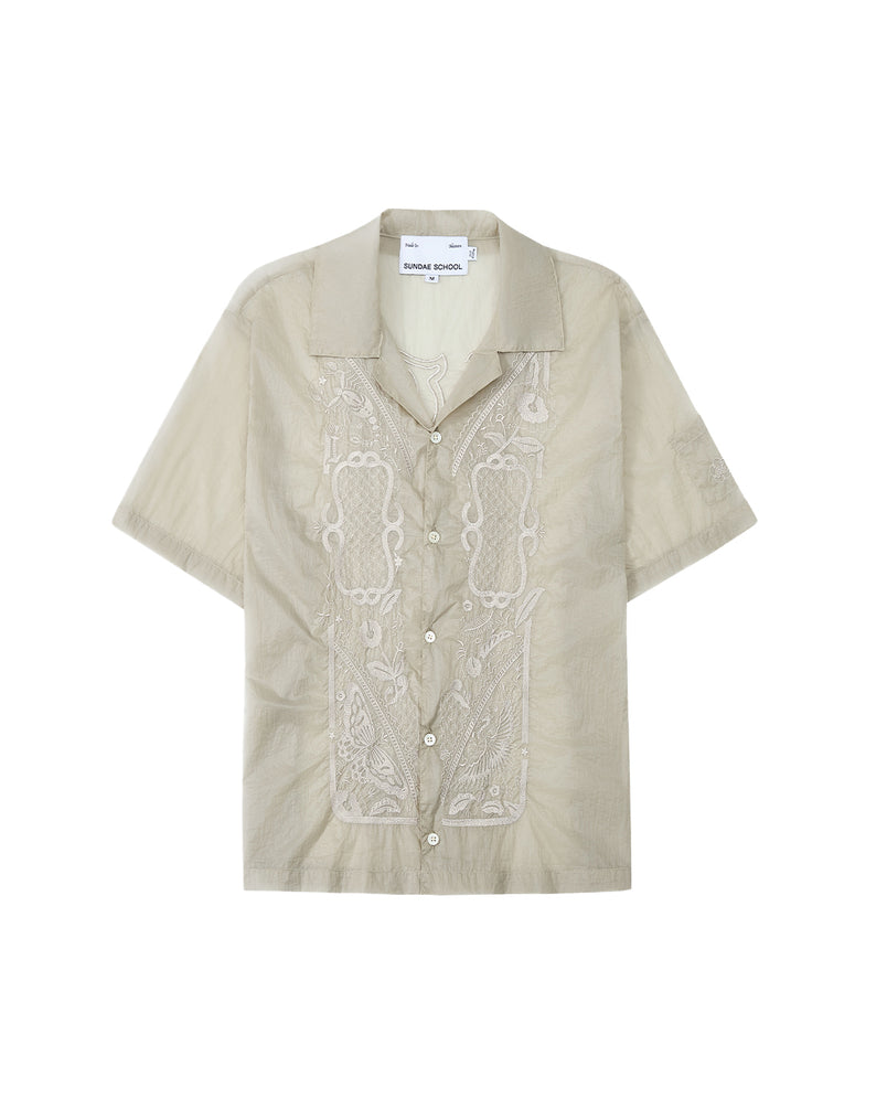 The BARONG SHIRT by Sundae School is a light beige short-sleeve button-up featuring intricate embroidery on the front, a camp collar, and design details inspired by the classic Filipino Barong Tagalog silhouette. This relaxed-fit shirt includes a visible Sundae School label inside the collar and subtle references to traditional craftsmanship.