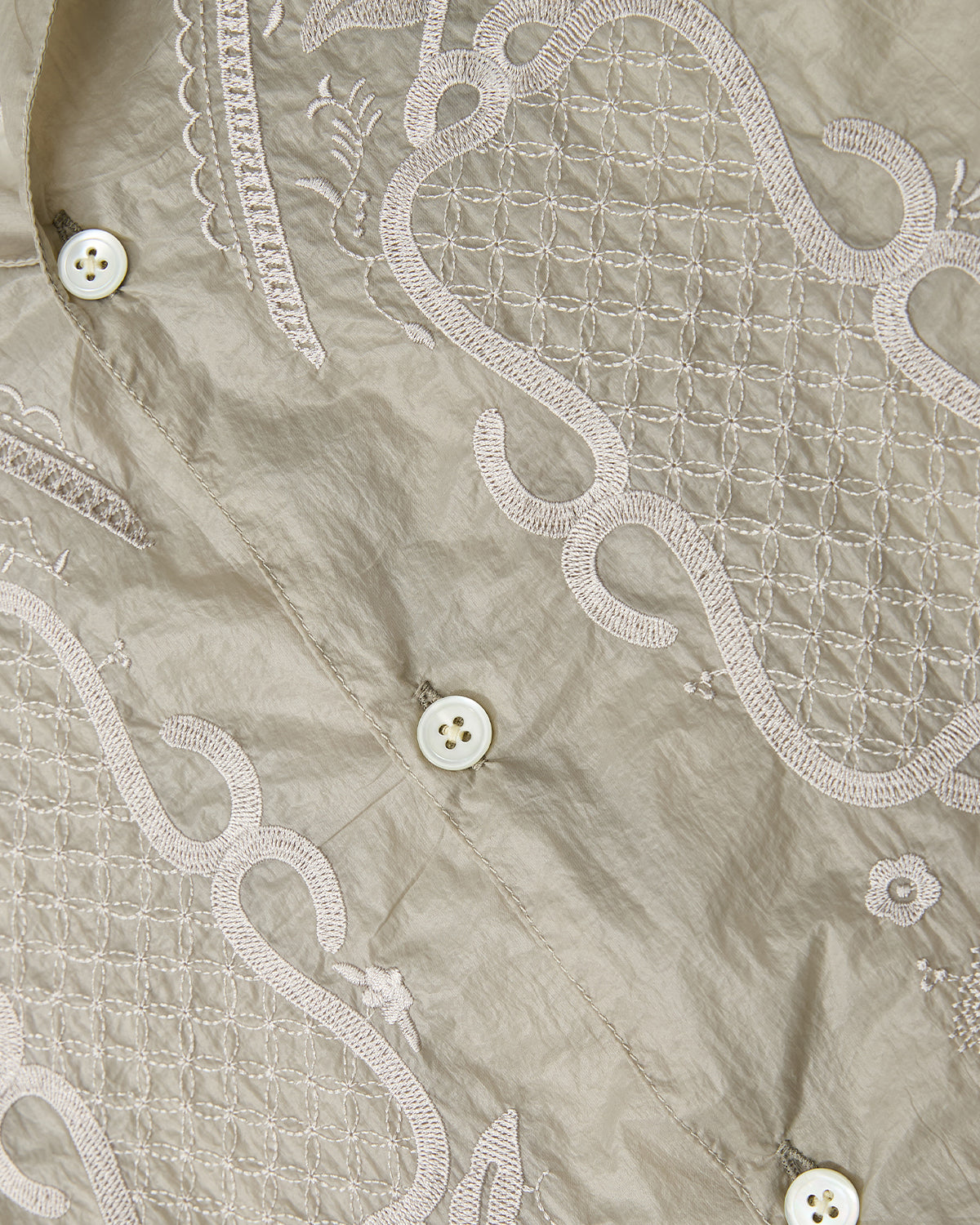 Close-up of the BARONG SHIRT by Sundae School, featuring beige fabric adorned with intricate embroidered patterns and white buttons sewn along the diagonal seam—evoking the timeless look of a traditional Barong Tagalog and showcasing the refined Filipino silhouette in a translucent nylon shirt.