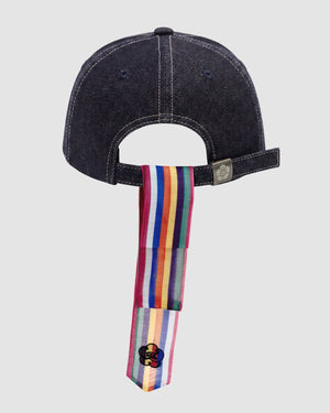 The INDIGO DAENGGI CAP by Sundae School is a dark denim, adjustable 6-panel cap with a strap featuring vertical rainbow stripes and a small black clover logo near the strap's end.