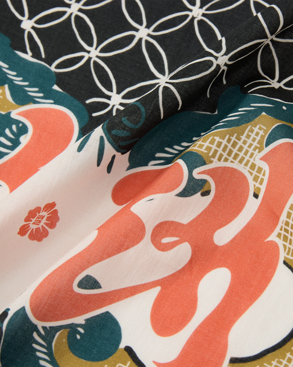 A close-up of the slightly wrinkled MULTI-COLOR BATIK BANDANA by Sundae School reveals abstract patterns—coral-red shapes, green accents, small flowers, and a black section with intersecting white circles—echoing Indonesian batik designs.