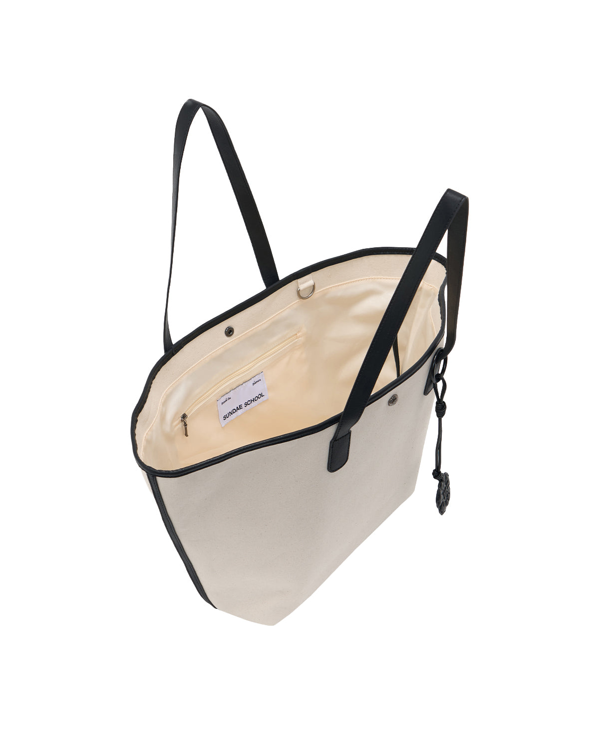 The IVORY BOJAGI TIE TOTE BAG by Sundae School features cream canvas with black vegan leather trim and handles. Shown open, it reveals a beige interior, a zippered pocket, and a small label inside. This chic and foldable tote is ideal for everyday use.