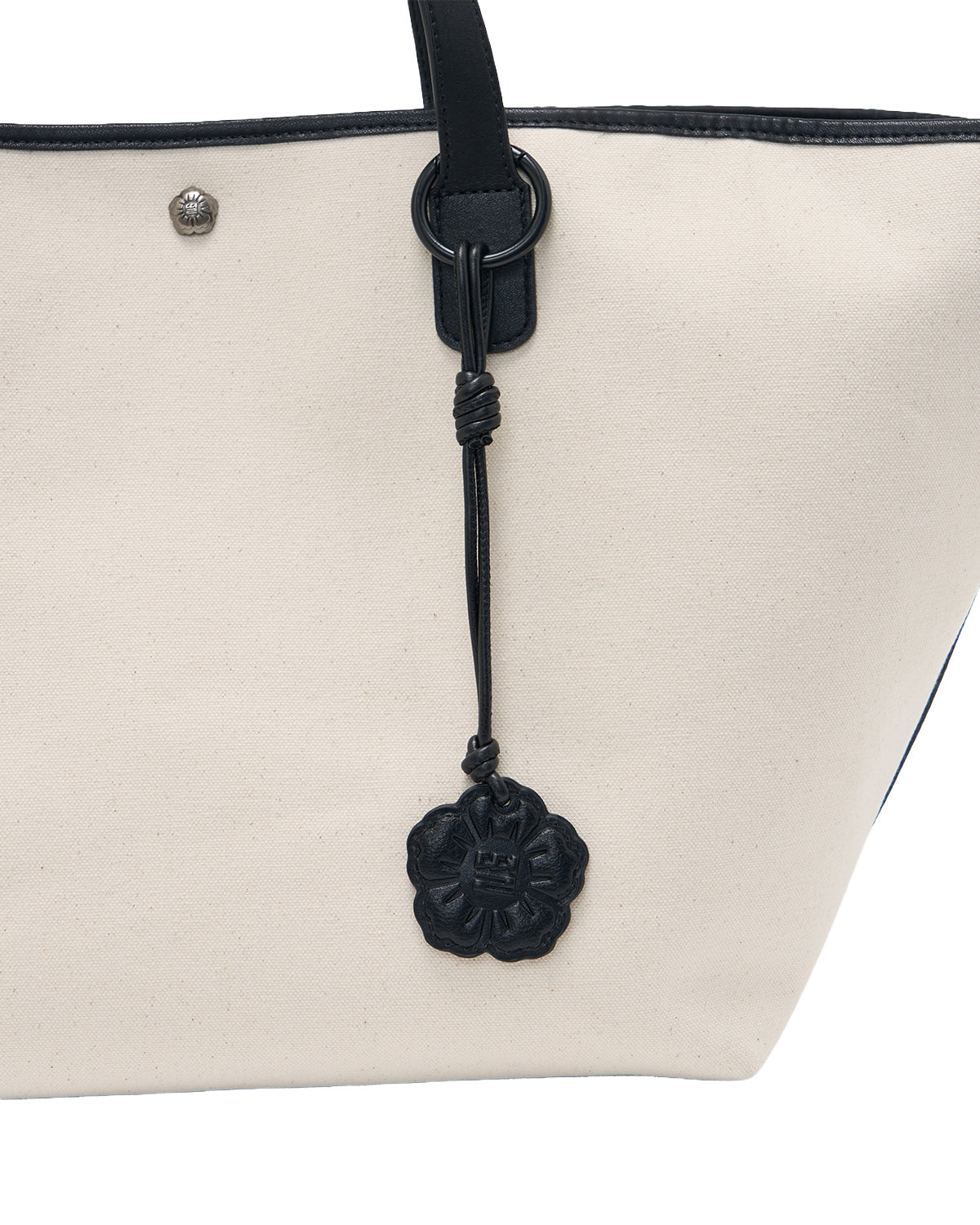 A close-up of the IVORY BOJAGI TIE TOTE BAG by Sundae School showcases its beige canvas body with vegan leather trim and black leather handles, complete with a striking black, floral-shaped leather charm attached to the strap. This stylish tote is both chic and practical—ideal for everyday use.