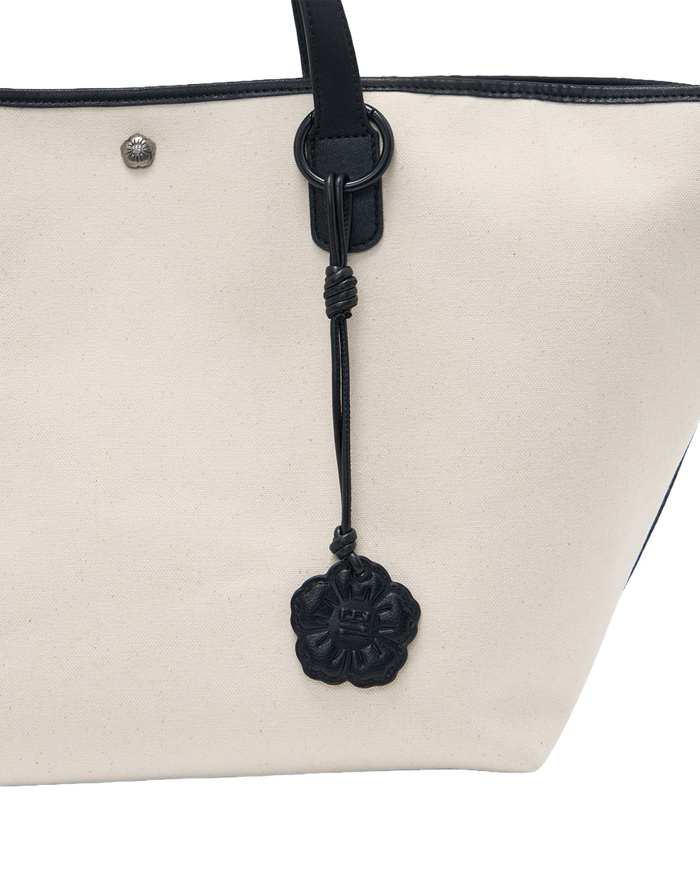 A close-up of the IVORY BOJAGI TIE TOTE BAG by Sundae School showcases its beige canvas body with vegan leather trim and black leather handles, complete with a striking black, floral-shaped leather charm attached to the strap. This stylish tote is both chic and practical—ideal for everyday use.