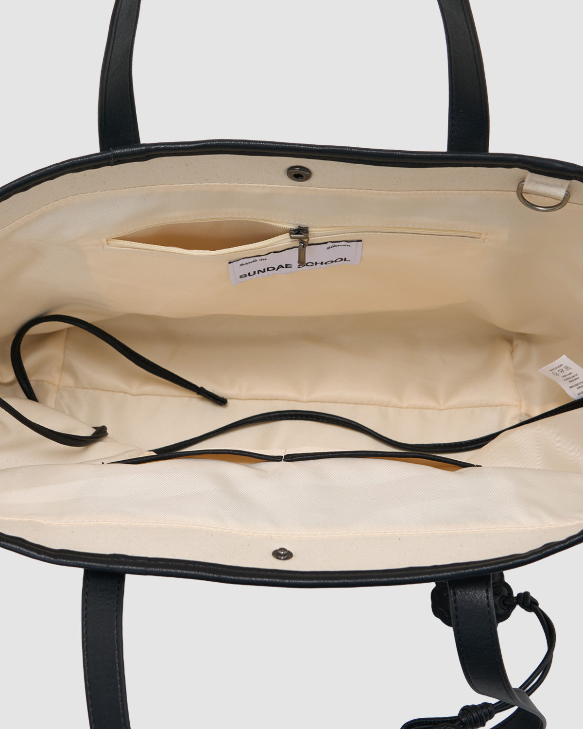 A close-up of the IVORY BOJAGI TIE TOTE BAG by Sundae School, featuring cream vegan leather with black trim, visible zippered and open pockets with a label, and black handles at the top.