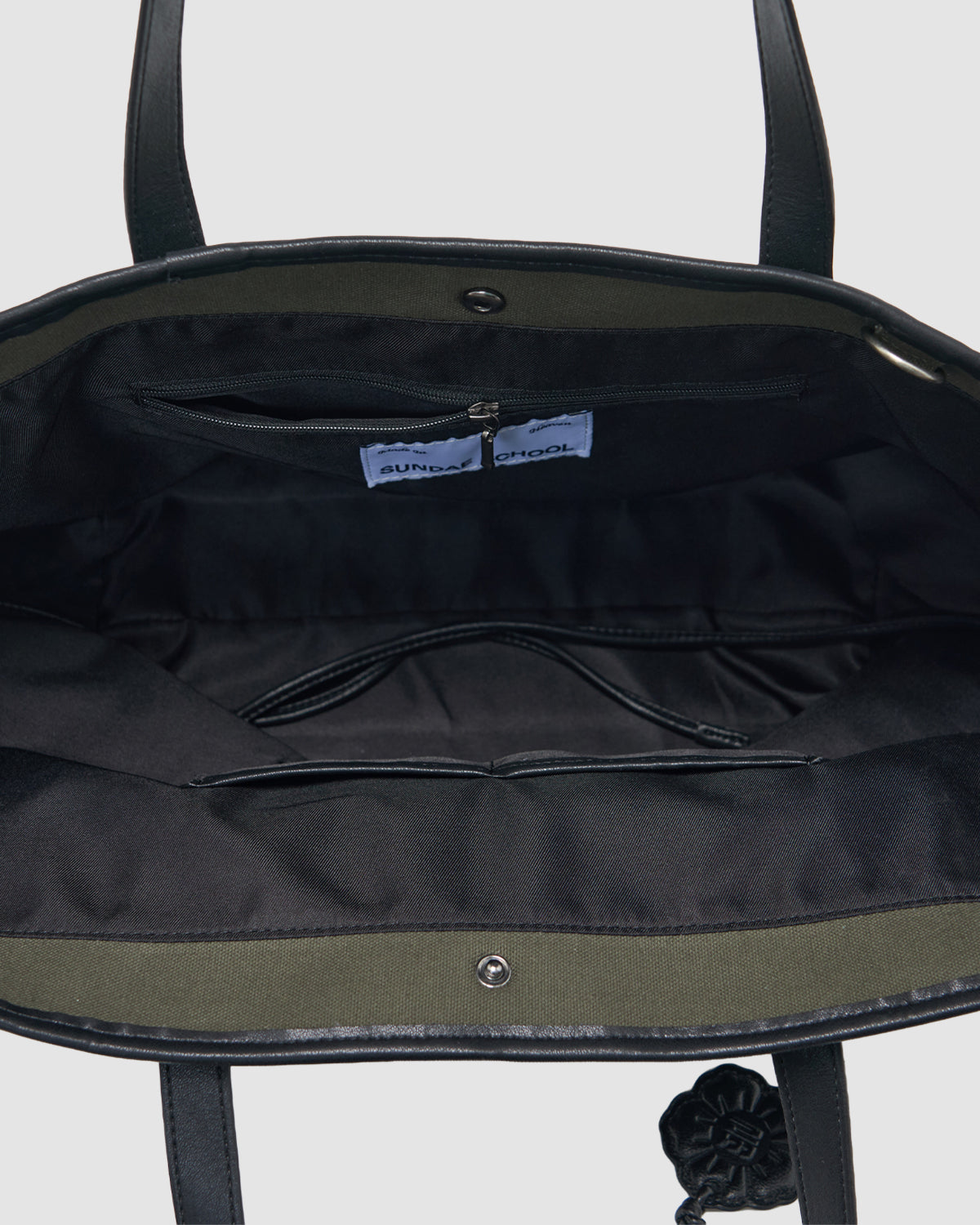 The OLIVE BOJAGI TIE TOTE BAG by Sundae School is a stylish, foldable canvas tote with an open black design, green inner lining, zippered pocket, fabric tags, vegan leather trim, two black handles, and a decorative black flower.
