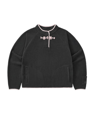 The CHARCOAL BUTTERFLY FLEECE PULLOVER by Sundae School is a black polar fleece sweater with light pink trim, featuring a mandarin collar adorned with decorative hand-tied flower knots at the neck. This mock-neck pullover offers two front pockets and a relaxed, casual fit.