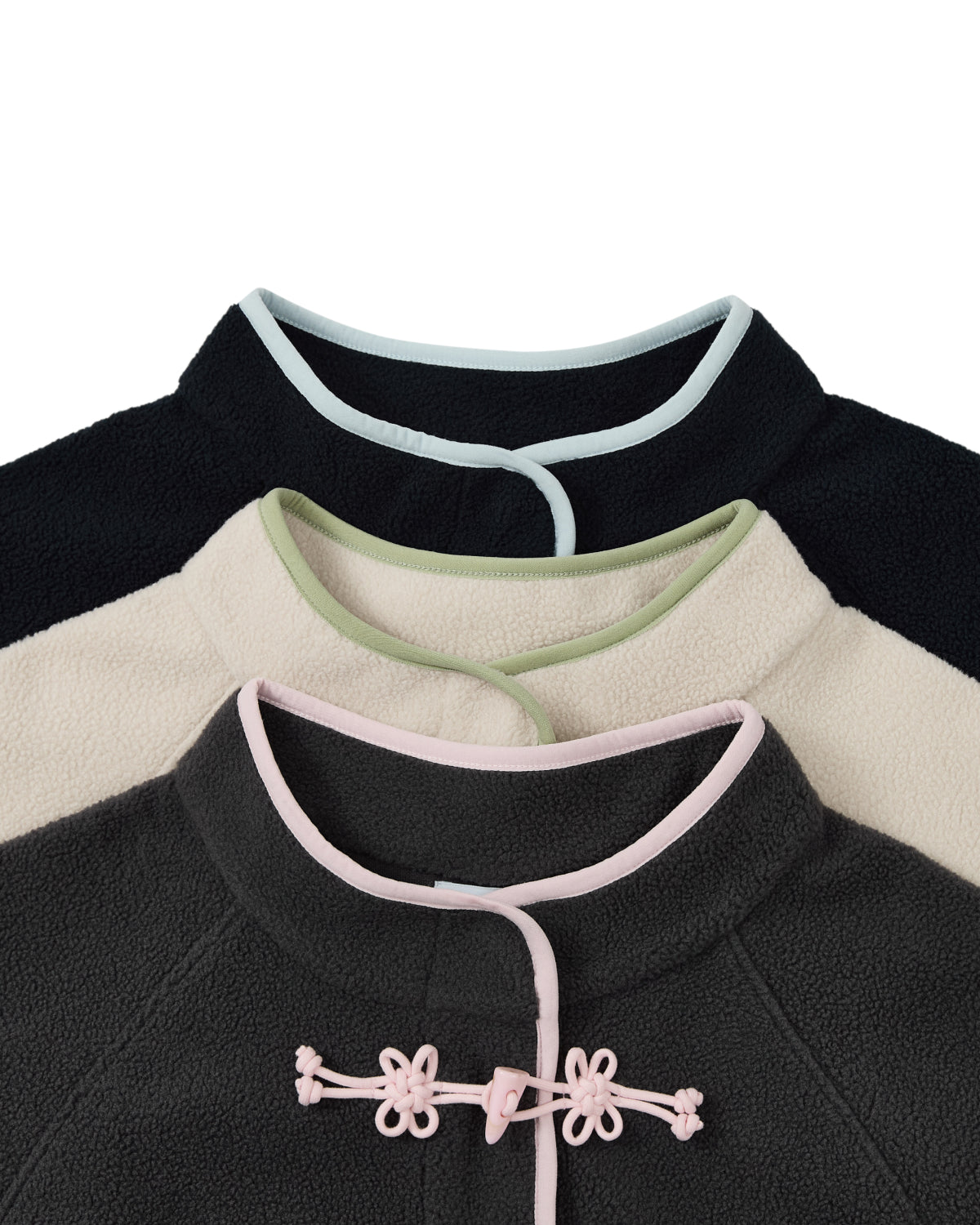 Three TAUPE BUTTERFLY FLEECE PULLOVERS by Sundae School are stacked, each adorned with a mandarin collar and contrast piping. The top pullover is a black mock-neck polar fleece with white trim, the middle features cream fabric with green trim, and the bottom is dark gray accented by pink trim and finished with hand-tied flower knots for added decoration.