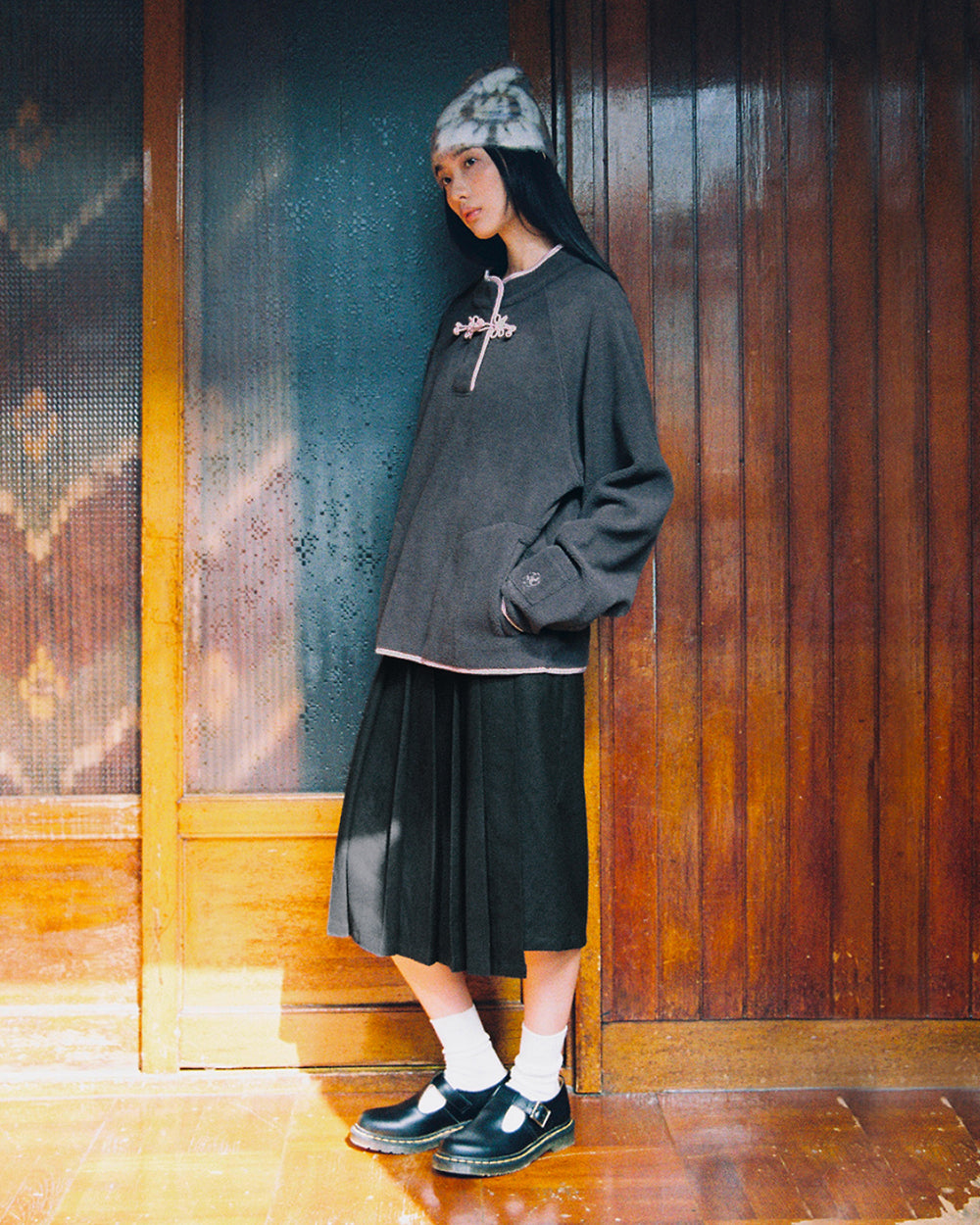 A person stands indoors wearing a furry hat, a loose dark CHARCOAL BUTTERFLY FLEECE PULLOVER from Sundae School, pleated skirt, white socks, and black shoes, with wooden paneling and sunlight in the background.